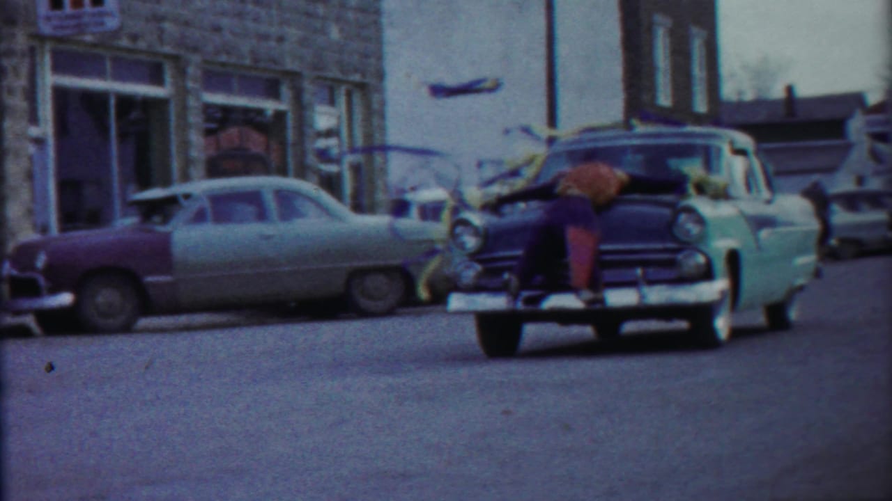 1958 Cheyenne County, Kansas home-movie frame showing a boy on the hood of a two-tone car near storefront windows.