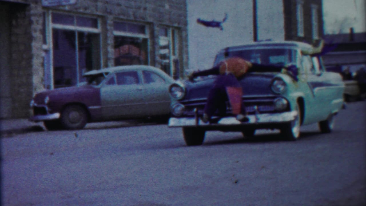 A two-tone car approaches with a boy stretched across its hood.