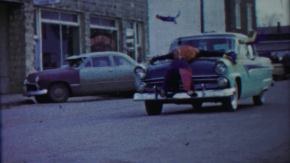 The car moves closer past stone storefronts, with the boy still lying across the front hood.