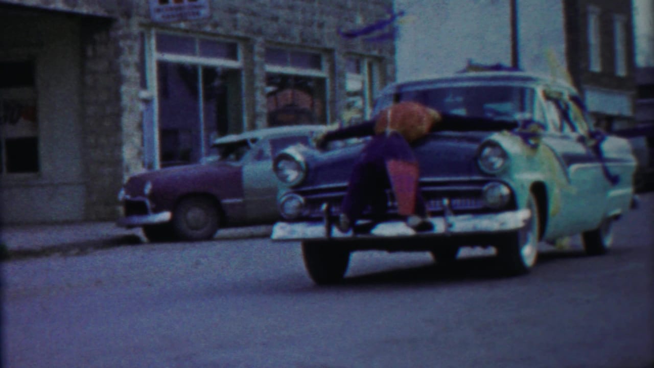 Home-movie frame from the Kansas street scene showing the boy on the hood beside a parked two-tone sedan and stone façade.