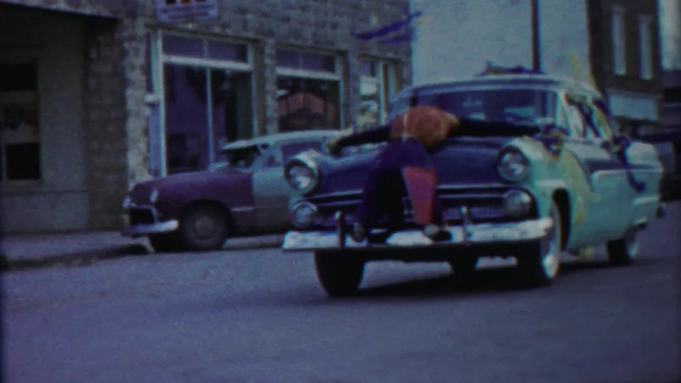 Home-movie frame from the Kansas street scene showing the boy on the hood beside a parked two-tone sedan and stone façade.