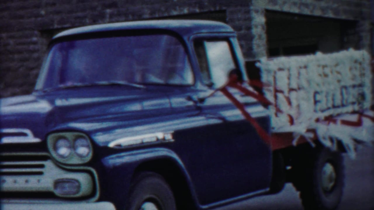 Close 1958 Kansas home-movie view of the pickup side, showing the cab, fender, red tie-downs, and stacked pale cargo.