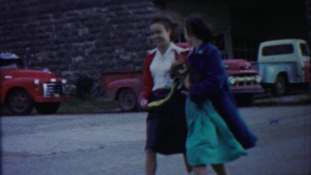 Two women walk past parked trucks near a stone building.