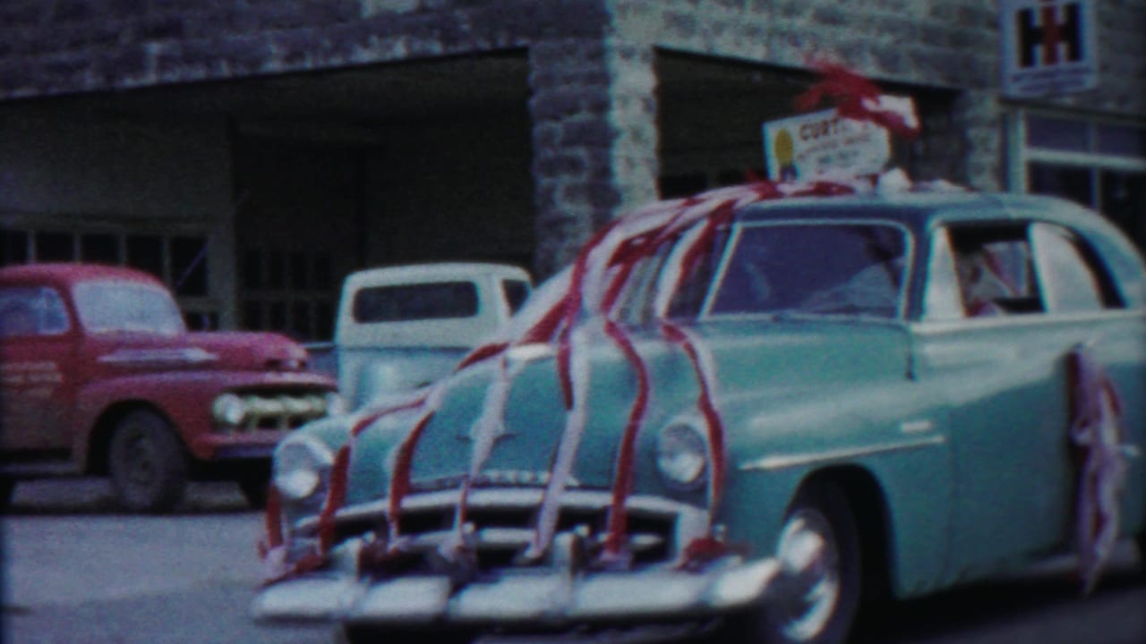 A light blue car covered in streamers drives past parked trucks.