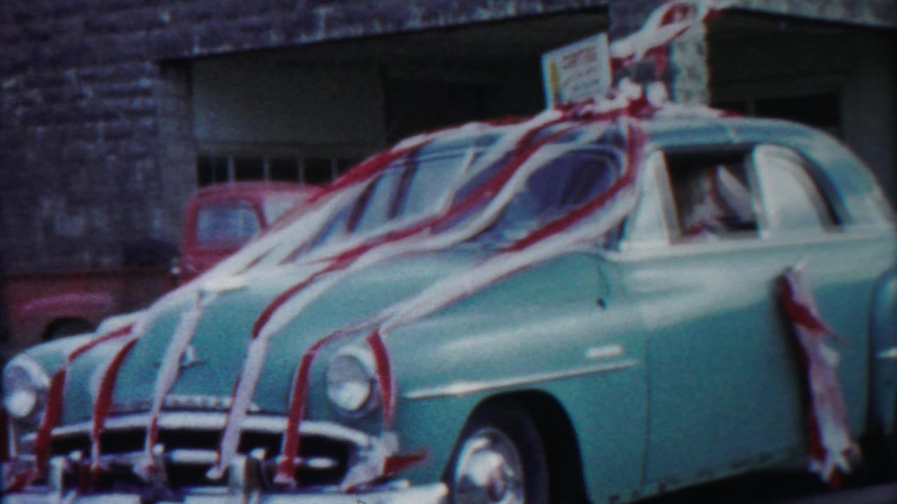 A decorated blue 1950s sedan fills the frame, with red and white streamers and a roof sign visible as it passes a stone building.