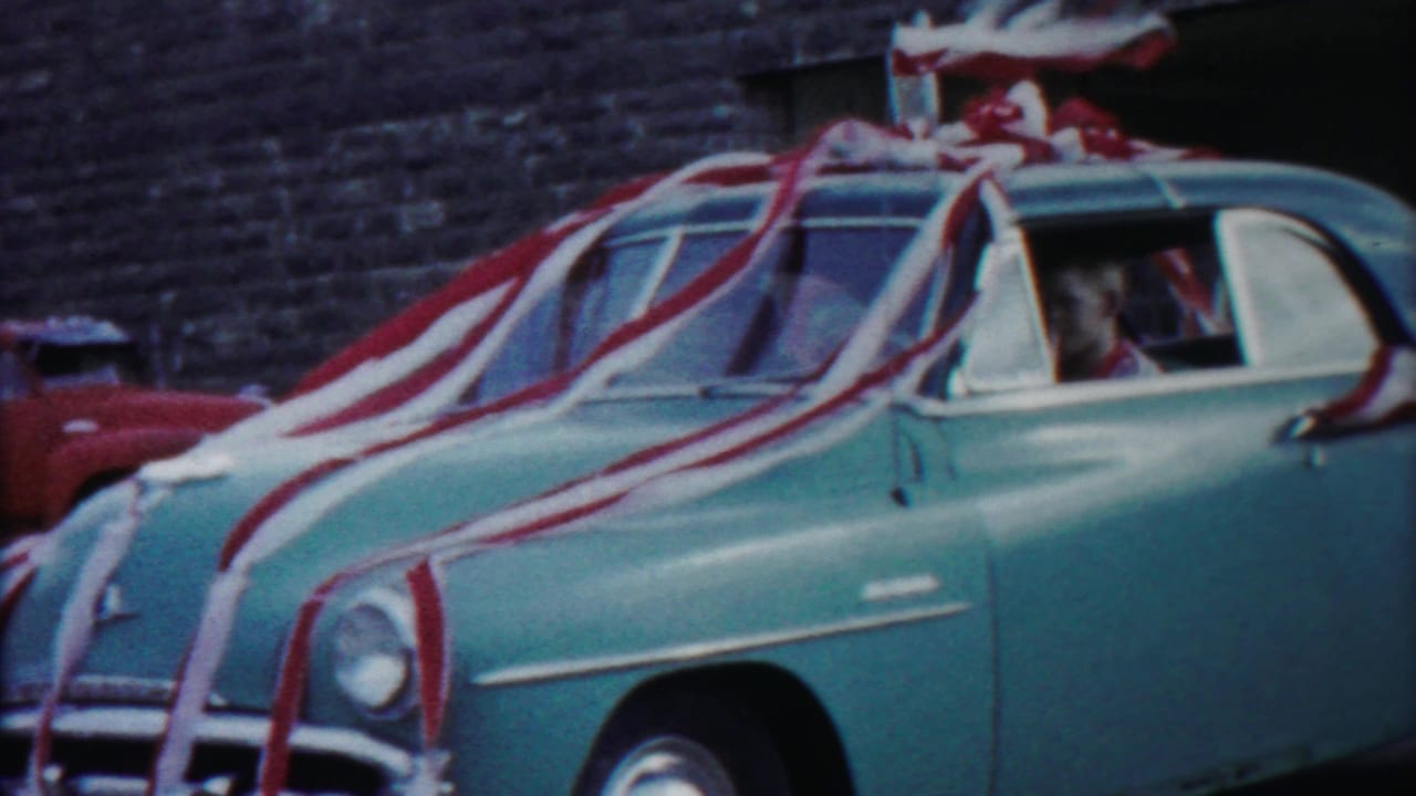 The blue decorated sedan continues past the camera, showing streamers draped across the hood and roof in a 1958 Kansas street scene.