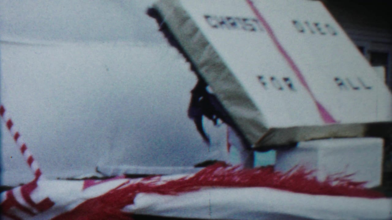 A church parade float in Kansas shows an open-book sign reading CHRIST DIED FOR ALL, with red trim and parked cars nearby.