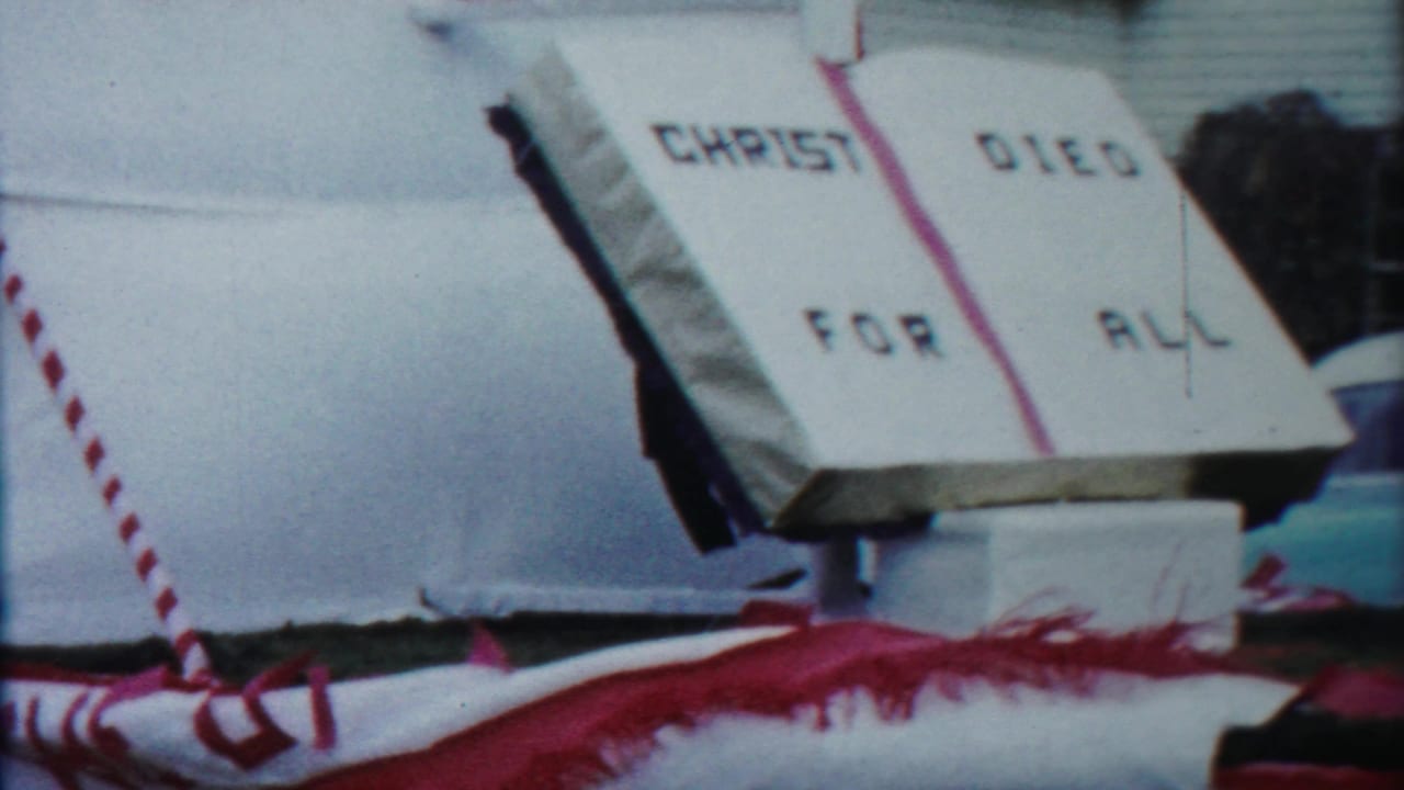 The 1958 home-movie view holds on the open-book church float sign, red fringe, striped support, and cars behind it.