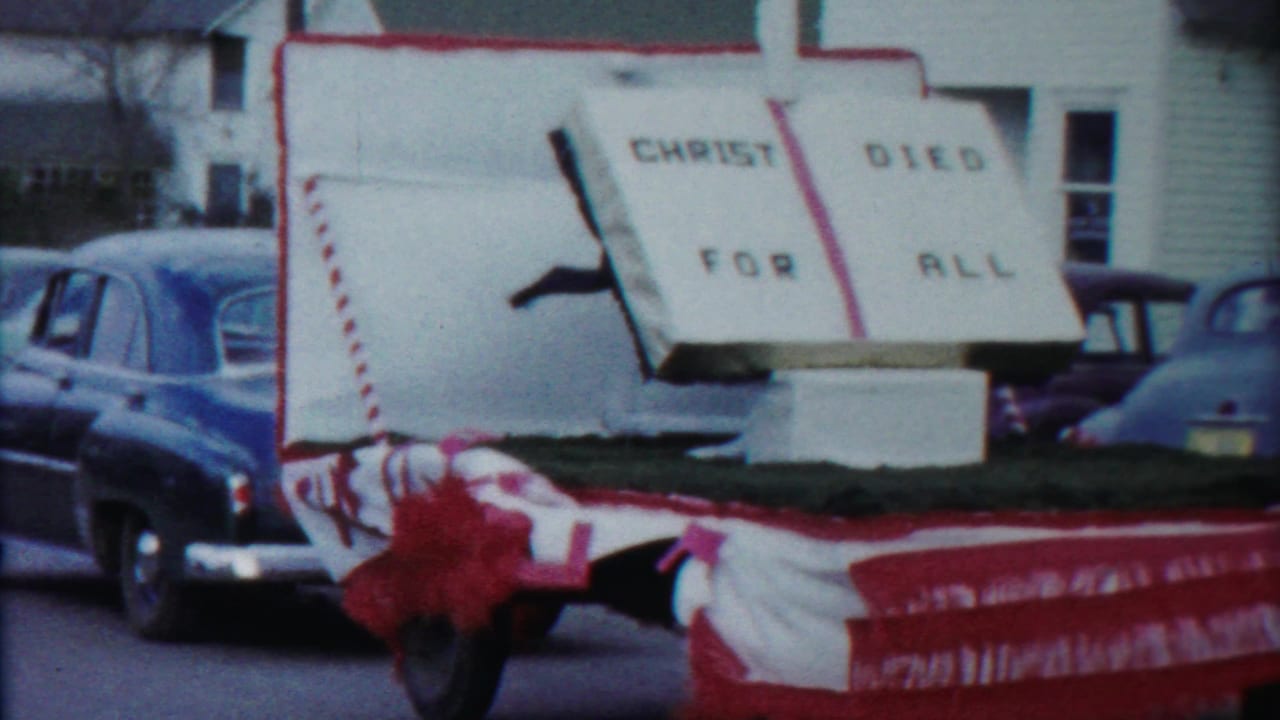 A Kansas parade float appears with a large open-book sign reading Christ Died For All, framed by parked 1950s automobiles.