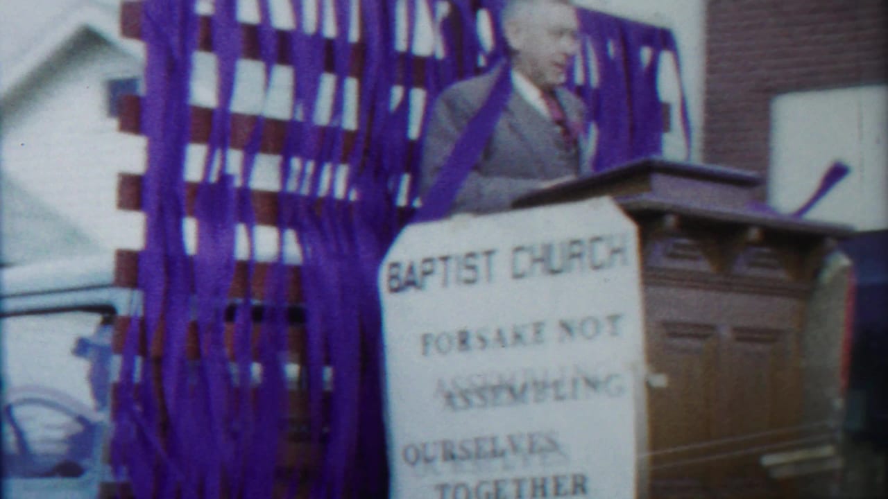 A suited speaker stands behind a wooden pulpit on a Baptist Church parade float decorated with purple streamers.