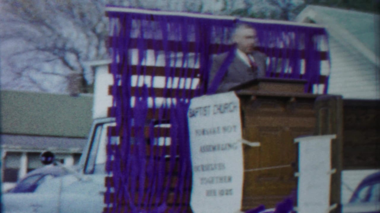 In Cheyenne County, Kansas, a suited speaker stands at a wooden podium on a Baptist church parade float decorated with purple streamers.