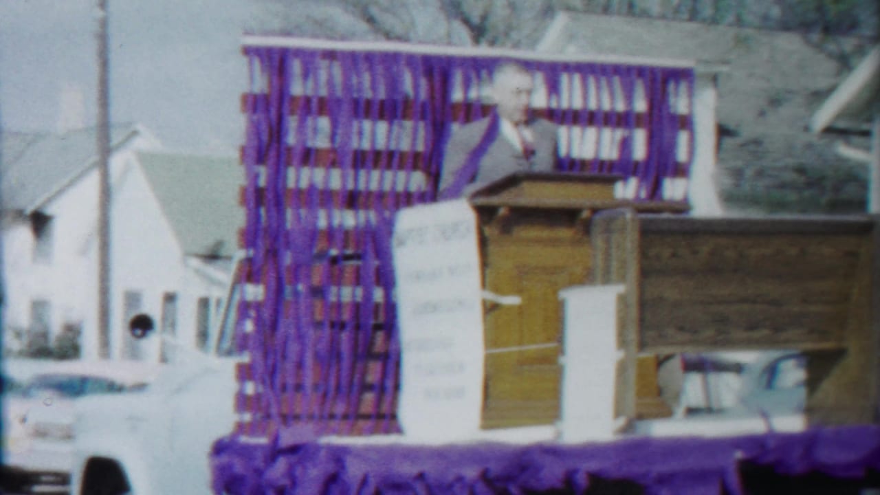 The Baptist church parade float moves along a residential street with purple streamers, a podium, a visible speaker, houses, and parked cars.