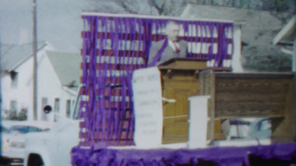 The Baptist church parade float moves along a residential street with purple streamers, a podium, a visible speaker, houses, and parked cars.