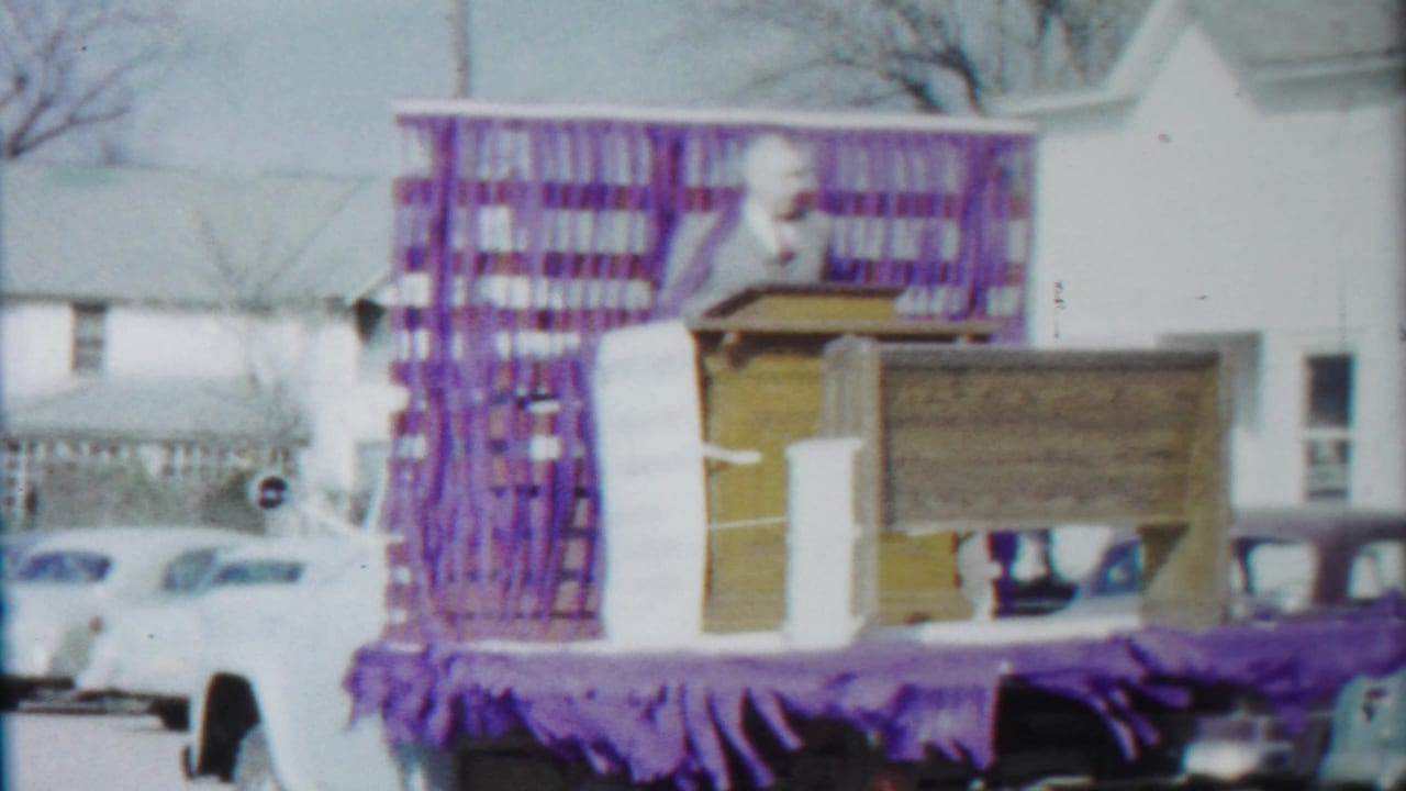 A side view shows the decorated Baptist church float, purple trim, wooden podium, and residential houses during the 1958 Kansas parade footage.