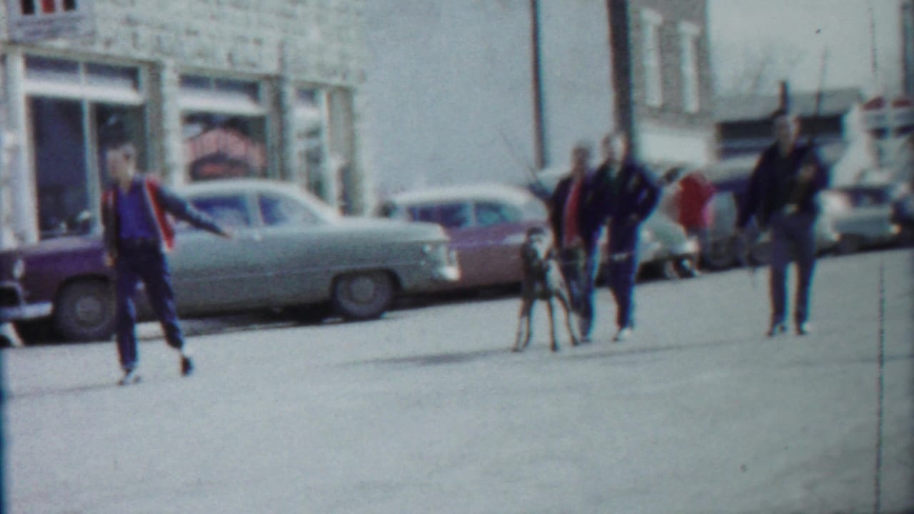 Young men and a large dog on a small-town street with parked vintage cars.