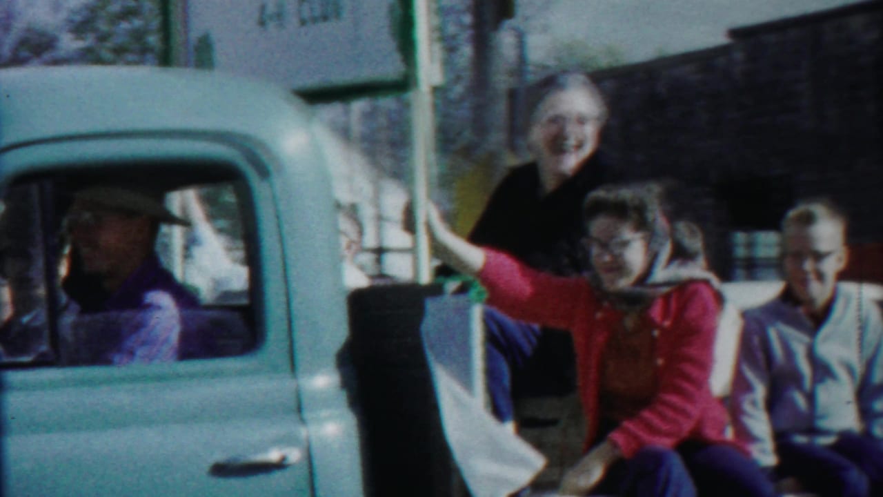Adults ride in the back of a pale blue pickup during the 1958 Cheyenne County parade, with one woman waving toward the camera.