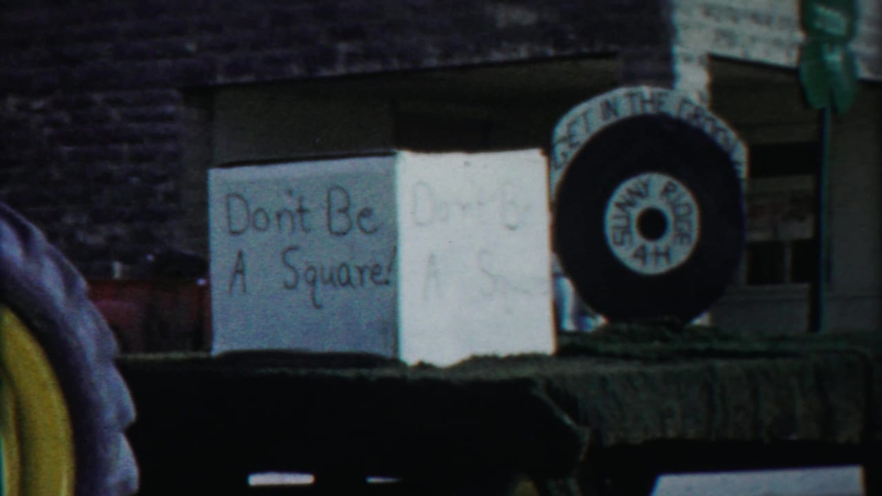 1958 Cheyenne County parade display with handmade “Don't Be A Square!” signage, a large tire, and a 4-H clover sign near brick storefronts.