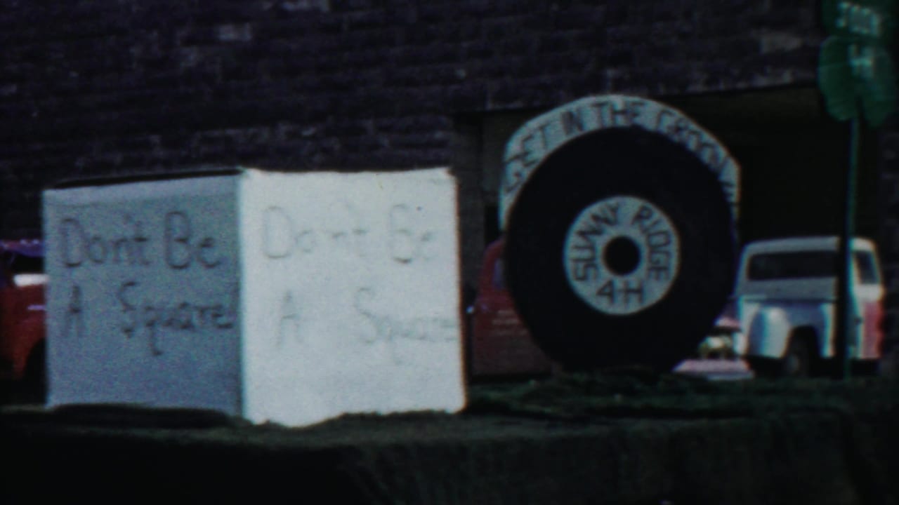 1958 Cheyenne County parade scene with handmade 4-H-themed signs, a large tire display, vintage vehicles, and a brick building in the background.