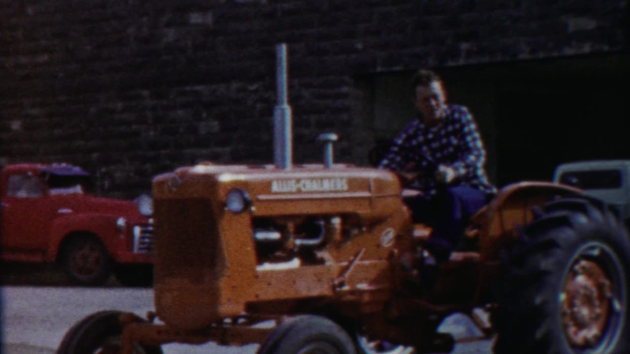 Man in a plaid shirt drives an orange Allis-Chalmers tractor in a 1958 Cheyenne County parade, with vintage vehicles behind him.