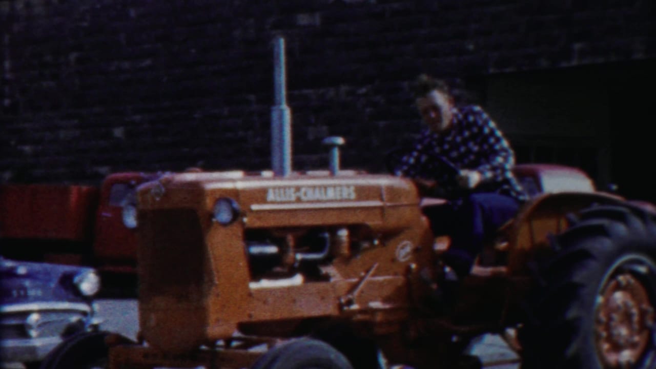 Man driving an orange Allis-Chalmers tractor past parked vehicles and a dark brick building.