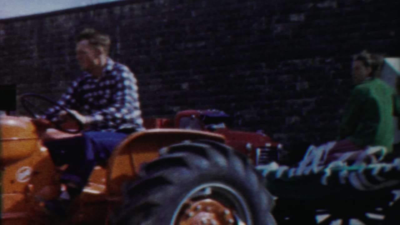 The orange tractor moves past as seated riders appear on a flatbed or trailer in the 1958 Kansas home-movie scene.