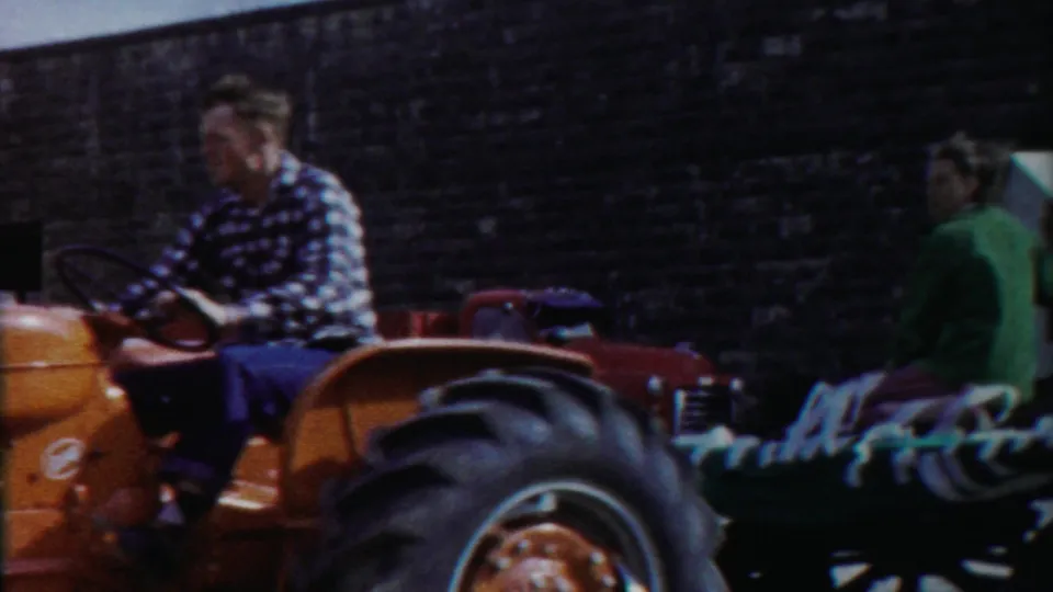 The orange tractor moves past as seated riders appear on a flatbed or trailer in the 1958 Kansas home-movie scene.