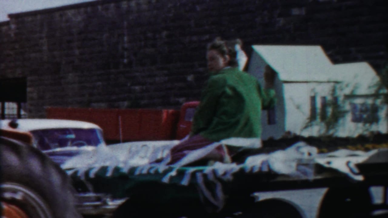 Two riders sit on a flatbed or trailer in Cheyenne County, Kansas, with parked vehicles and farmyard buildings in view.