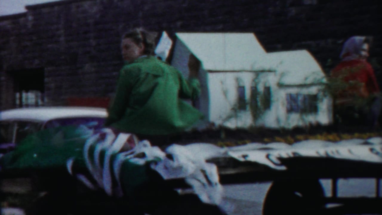 Decorated parade float with seated riders and a miniature white house passing a stone building.