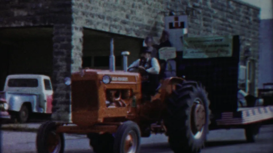 Red Allis-Chalmers tractor appears to pull a decorated parade platform past parked vehicles in Cheyenne County Kansas, 1958.