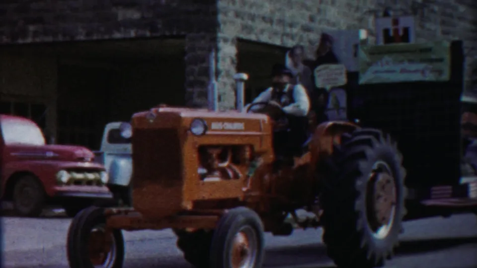 The tractor-drawn float continues past a stone storefront and parked mid-century vehicles during the 1958 Kansas parade.