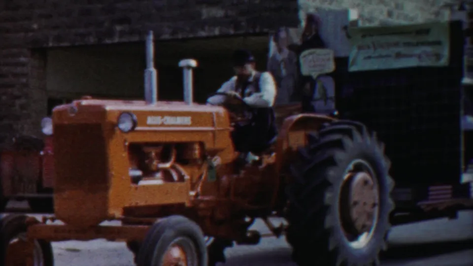 Orange tractor with visible branding moves through the Kansas parade with a costumed rider on the float behind it.