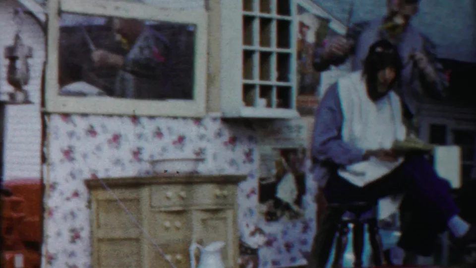 The parade float presents a staged barbershop scene with a seated performer, chair, wallpaper panels, and painted props.