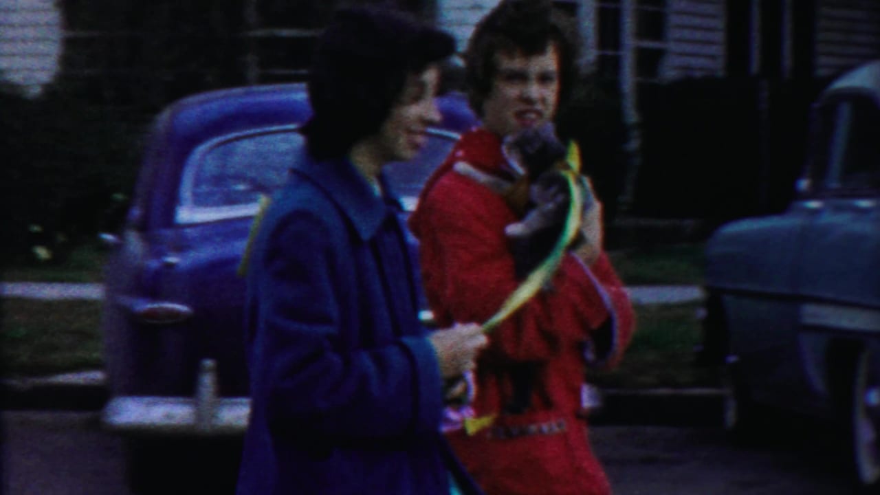Two women walk past vintage cars in a residential street parade.