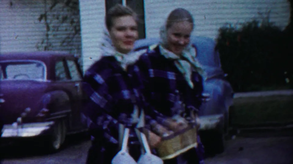 Two women, one in plaid outerwear and one in a headscarf, carry baskets while walking along a residential street in the Kansas parade footage.