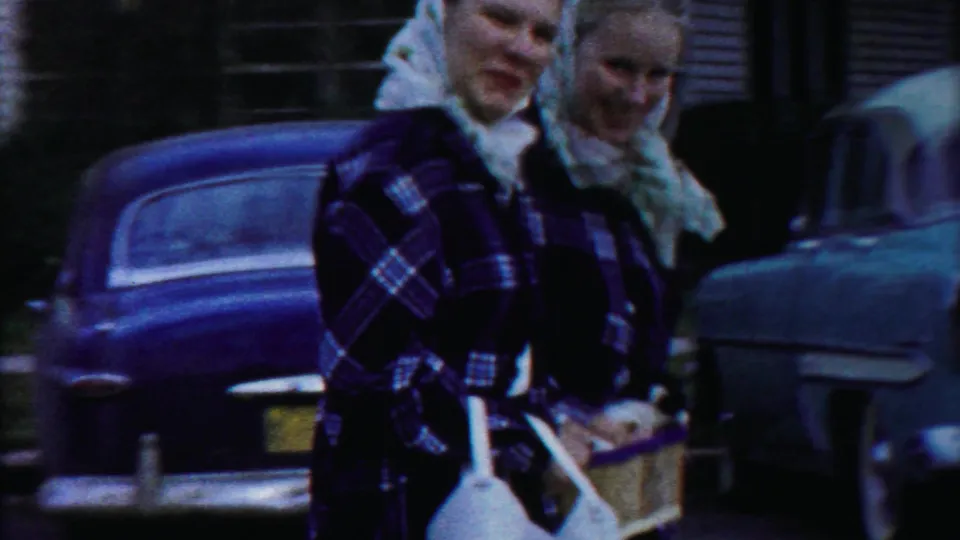 The parade participants pass close to the camera, with baskets, a white prop, and parked 1950s cars visible behind them.