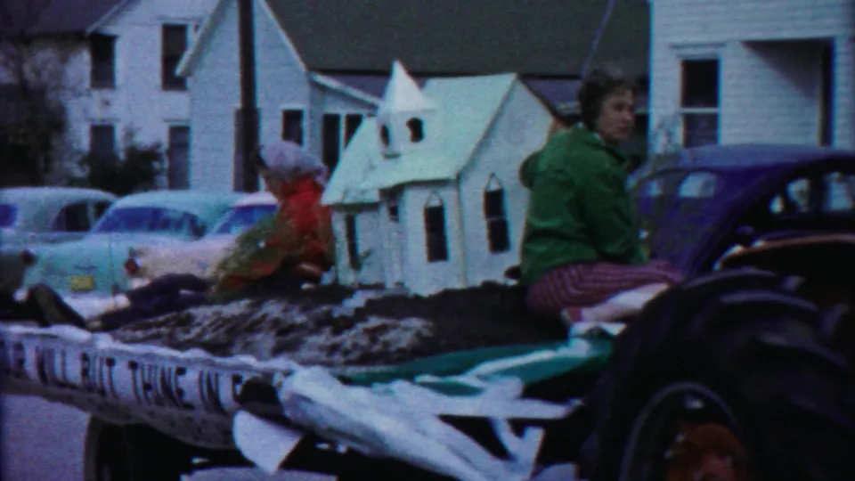 A float carrying a small white building model moves through a residential Kansas street, with vintage cars and houses in the background.