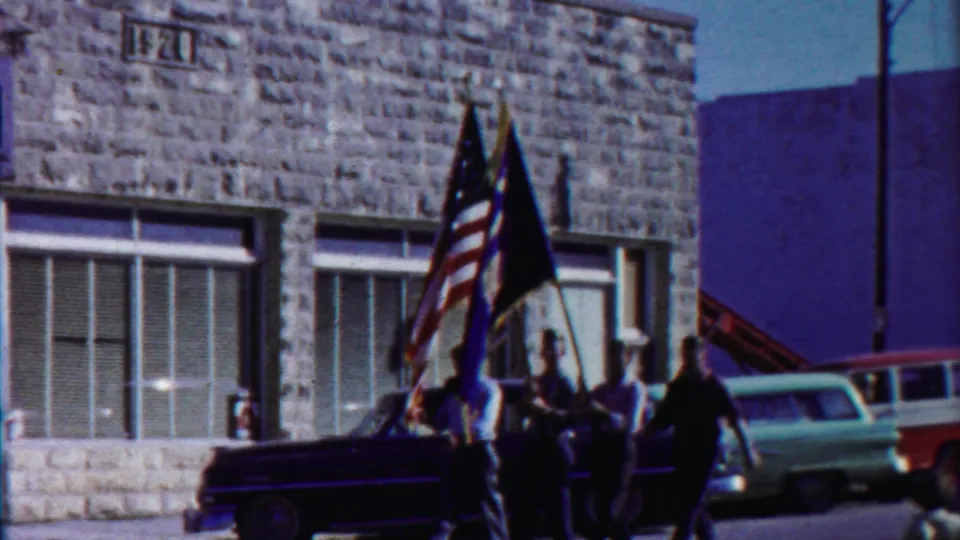 Parade participants hold American and other flags beside a dark car outside a stone-front building with an IH sign in Cheyenne County.