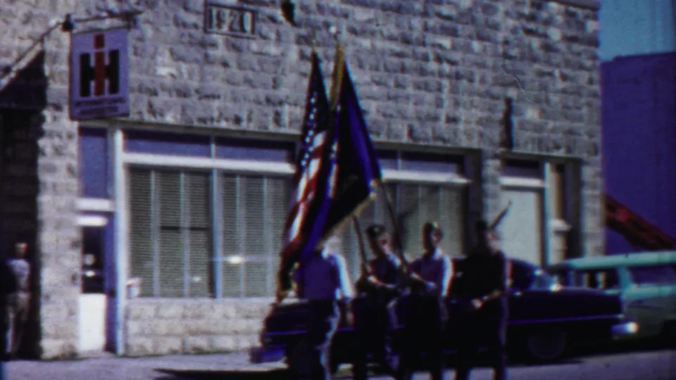 Parade flag bearers stand beside a dark car near a stone storefront with an IH sign, with mid-century vehicles parked along the street.