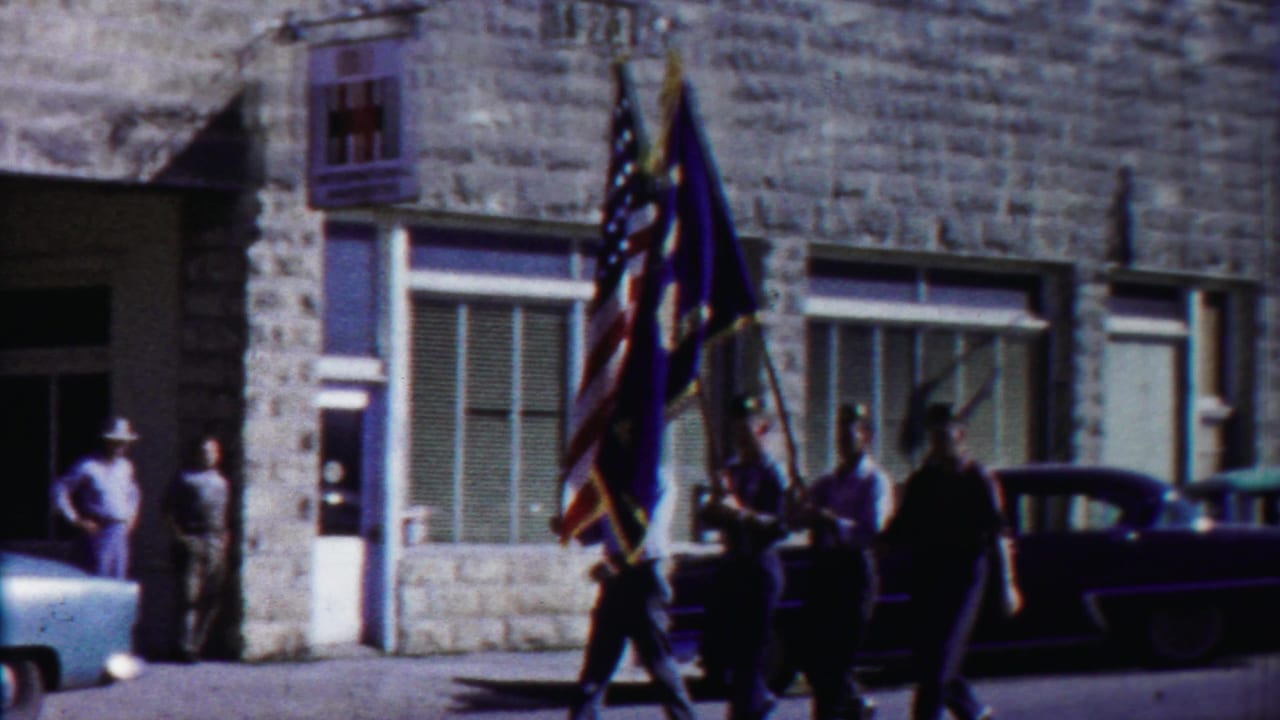 1958 Cheyenne County, Kansas parade view with young men carrying flags past stone storefronts and mid-century parked cars.