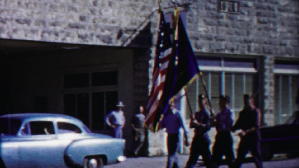 Patriotic parade participants carry an American flag and dark banner past a light blue sedan in Cheyenne County, Kansas.