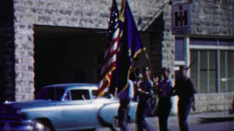 Home-movie frame of flag bearers in a 1958 Kansas parade moving beside a light blue vintage car and storefront windows.