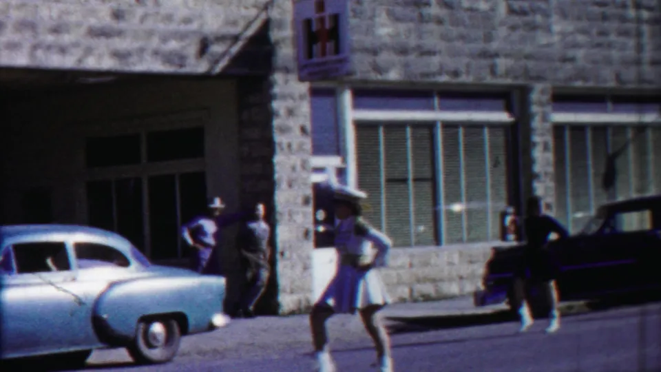 Street-level 1958 Cheyenne County parade view with a woman in a light dress, a parked car, storefront windows, and spectators.