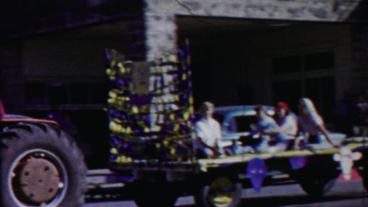 Decorated parade float passing a stone building with riders and a large tractor wheel nearby.