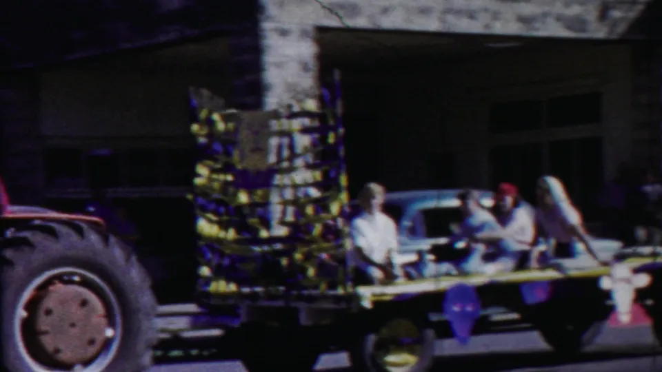 1958 Kansas home-movie frame of a decorated float moving past a stone building, with riders in period clothing and a tractor wheel at the edge.