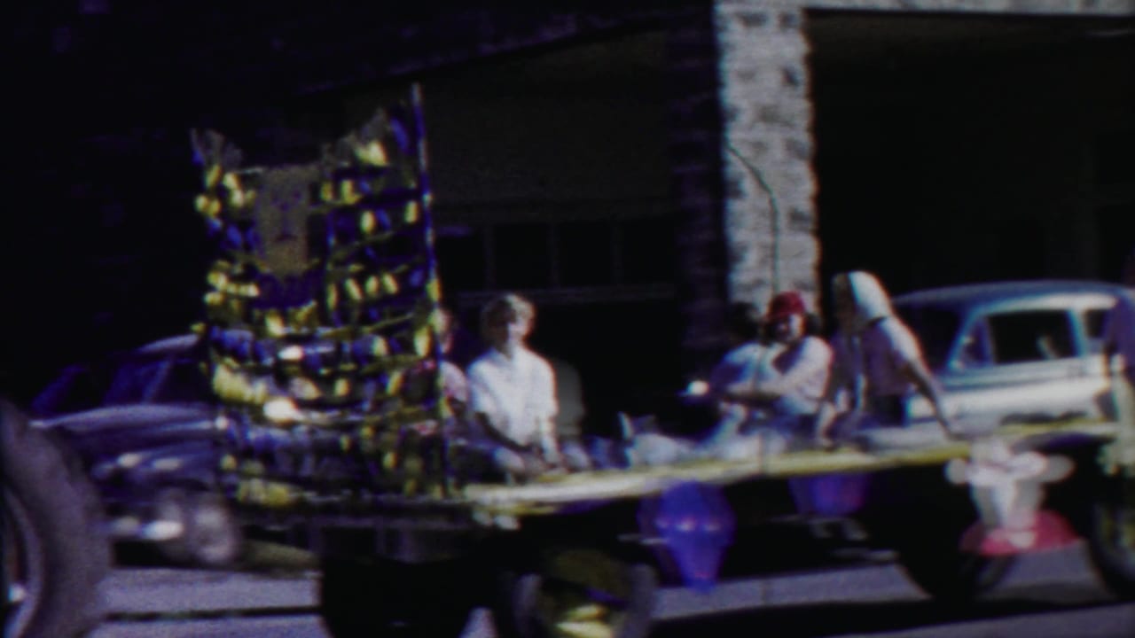Parade float with seated and standing riders moving past parked mid-century cars.