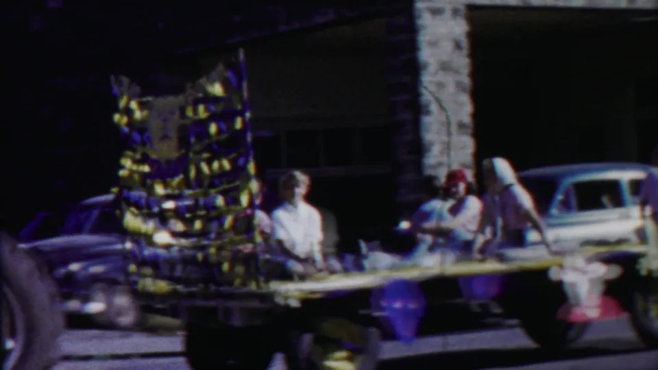 1958 Kansas parade frame showing riders on a decorated float, parked cars, and a stone-front building in the background.