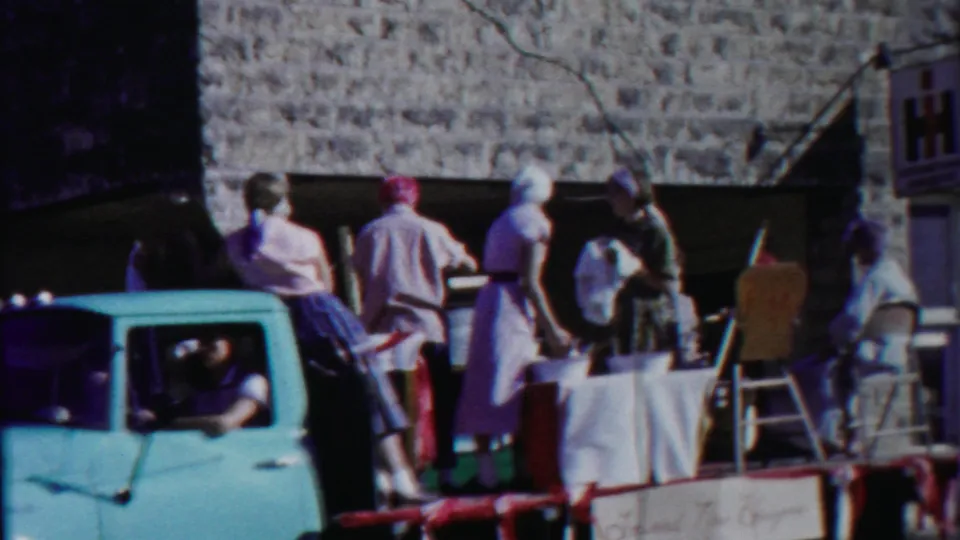 1958 Kansas home-movie frame with a light blue pickup cab, float participants in period clothing, chairs, a table, and a stone building behind them.