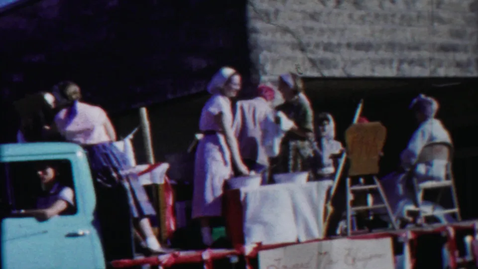 1958 Kansas parade frame showing women in aprons and headscarves on a float with chairs, a table, and a light blue pickup in front.