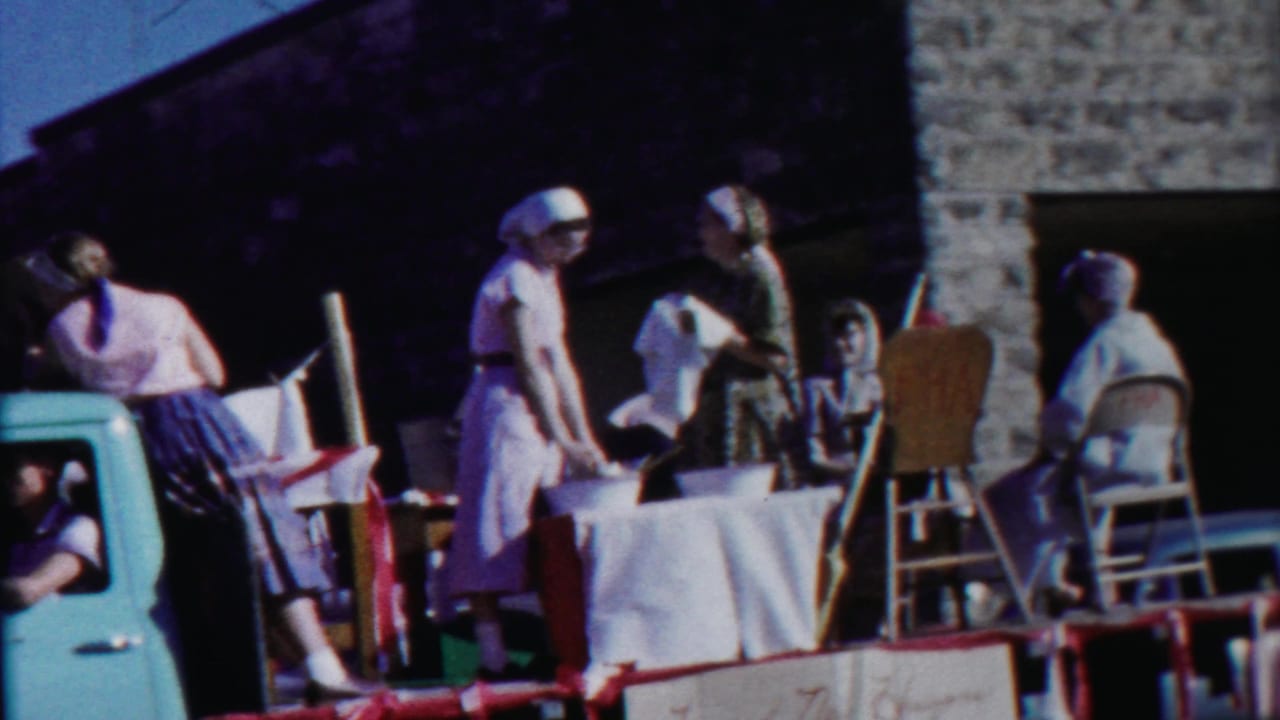 Women stand on a parade float beside a white-covered table and chairs near a light blue truck cab.