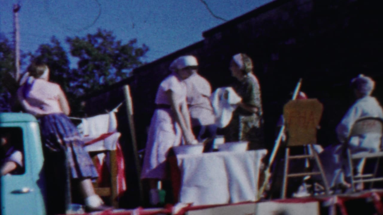 Parade float with women around a table, chairs, and a dark building edge behind them.